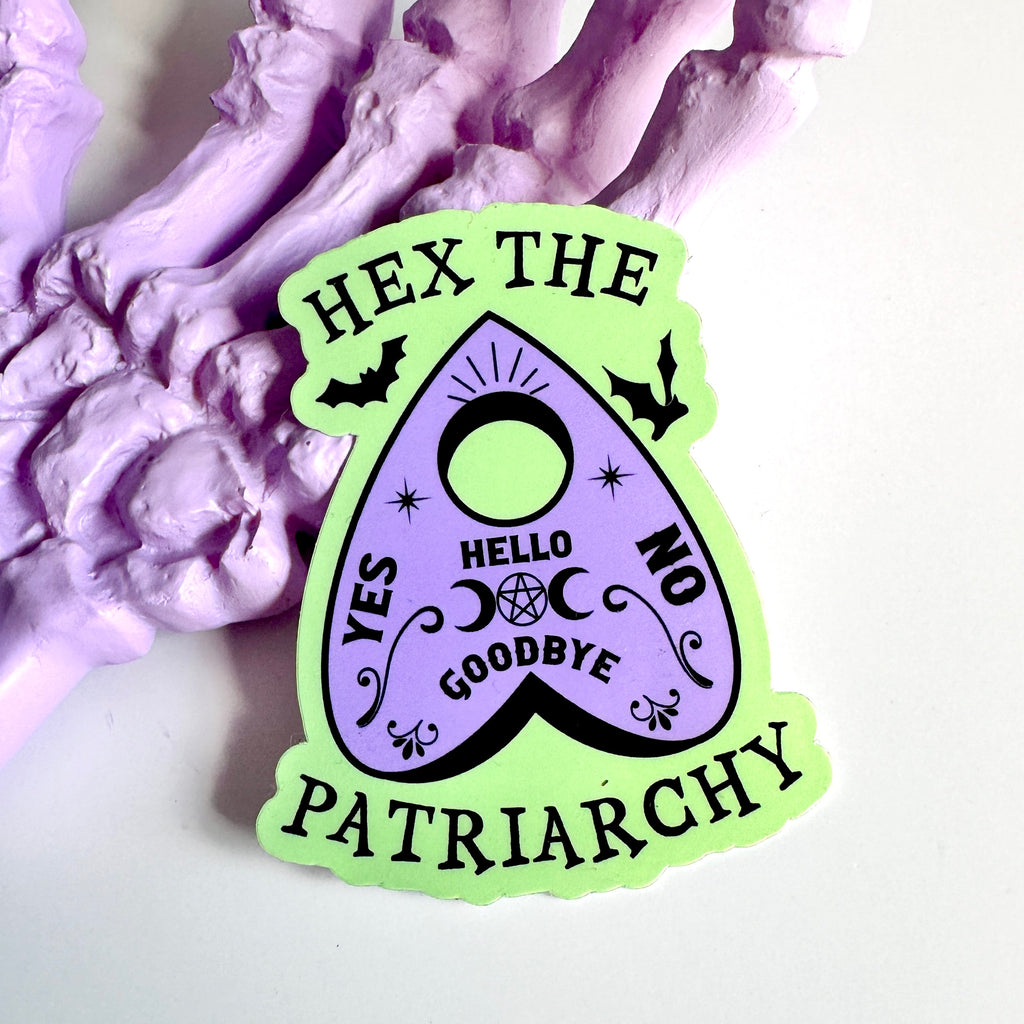A pastel green and lavender planchette-shaped sticker with black text that reads ‘Hex the Patriarchy.’ The design features bats, stars, moons, and the words yes, no, hello, and goodbye in a witchy spirit-board style. The sticker is placed on a lavender-painted skeleton hand.