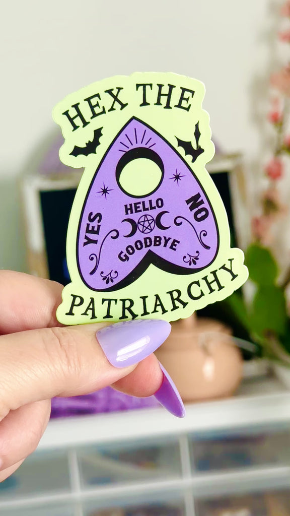 Hex the Patriarchy witchy planchette sticker in pastel green and lavender with bats, moons, and stars, feminist witchcraft laptop decal resting on a lavender skeleton hand.