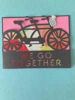 Custom cut greeting card in gray featuring a craft paper tandem bike with patterned paper backing over the words We Go Together in a horizontal opening. Inside is a pink layered paper over the charcoal grey outer card to write your note in. 