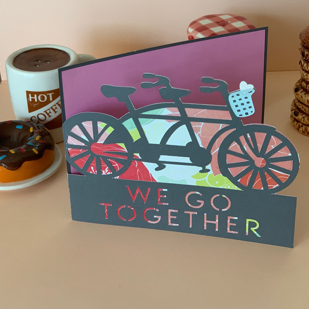 
            
                Load image into Gallery viewer, Custom cut greeting card in gray featuring a craft paper tandem bike with patterned paper backing over the words We Go Together in a horizontal opening. Inside is a pink layered paper over the charcoal grey outer card to write your note in. 
            
        